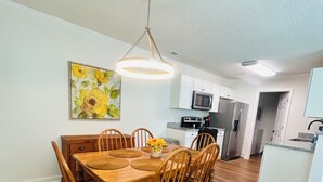 A dining area with a wooden table and chairs, a hanging light fixture, a kitchen in the background, and a painting on the wall.