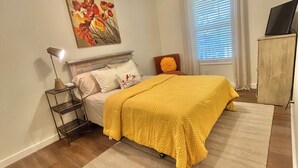 A bedroom with a bed, a ceiling fan, a painting, a window with blinds, a nightstand, and a TV on a cabinet.