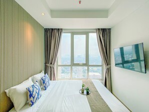 A hotel room with a large bed, a flat-screen TV on the wall, and a city view through the window. - Bright And Spacious Studio Apartment At Antasari Place (Jakarta)