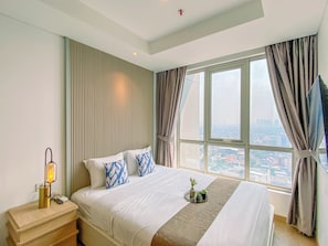 A hotel room with a large bed, a bedside table with a lamp, a view of the city through the window, and a curtain. - Bright And Spacious Studio Apartment At Antasari Place (Jakarta)