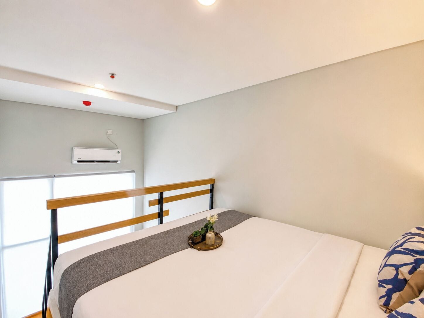 A bedroom with a bed, a window, an air conditioning unit, and a small plant on the bedside table.