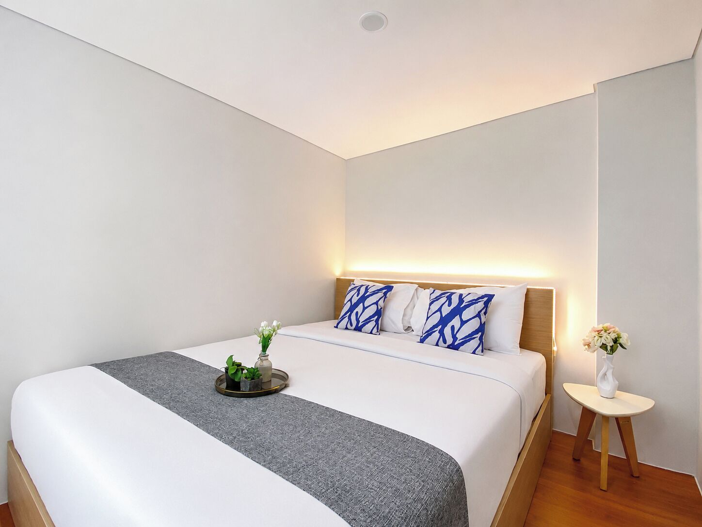 A modern hotel room with a large bed, a bedside table with a vase, and a wall-mounted light.