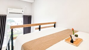 A hotel room with a bed, a window with curtains, an air conditioning unit, and a small table with a plant and a vase. - Urban Cozy Loft Studio At Apartment Apple 3 Condovilla (Jakarta)