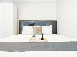 Una habitación de hotel moderna con una cama grande, ropa de cama blanca, un cabecero gris y una bandeja con un florero, una planta pequeña y una vela.