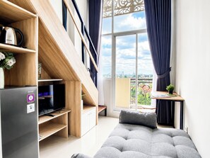 A modern apartment with a grey sofa, a Sharp refrigerator, a wooden shelving unit, and a balcony with a view of the city.