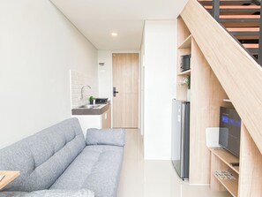 Un espai habitable petit amb un sofà gris, una cuina amb un rentapeus i un frigorífic, i unes escales de fusta.