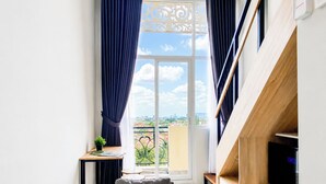 A modern room with a sofa, a small table with a plant, a TV, and a view of the city through a large window with curtains. - Cozy Design Studio Loft At Apple 3 Condovilla Apartment (Jakarta)
