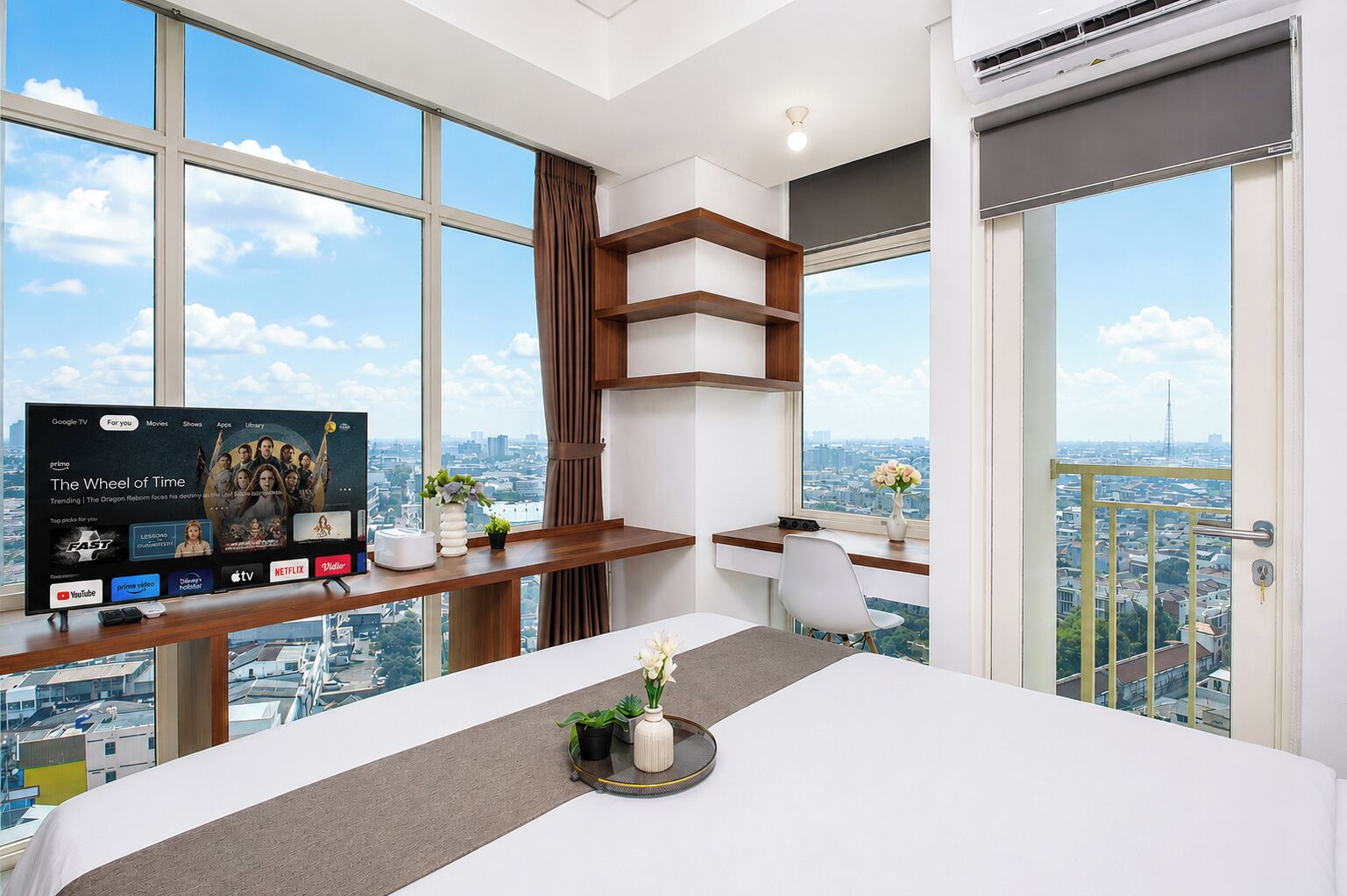 A modern hotel room with a large TV, a desk with a computer, a bed, and a balcony with a city view.