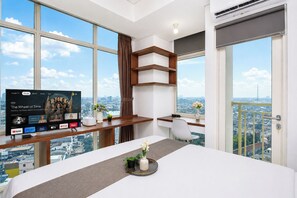 A modern hotel room with a large TV, a desk with a computer, a bed, and a balcony with a city view. - Cozy Studio With City View Apartment At B Residence Grogol (Jakarta)