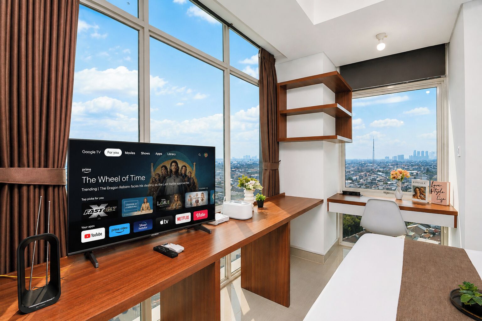 A modern office space with a large flat-screen TV displaying a streaming service interface, a wooden desk with a smartphone, and a city view through large windows.