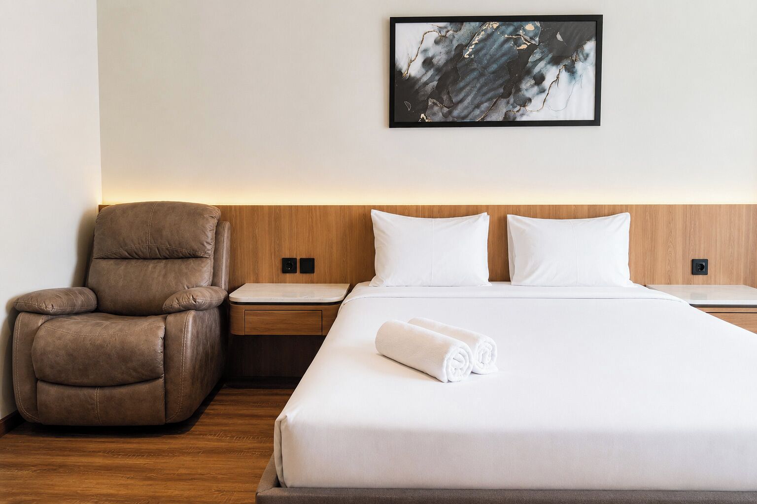 Une chambre d’hôtel avec un lit, un fauteuil en cuir marron, une table de chevet et une œuvre d’art abstraite encadrée accrochée au mur.