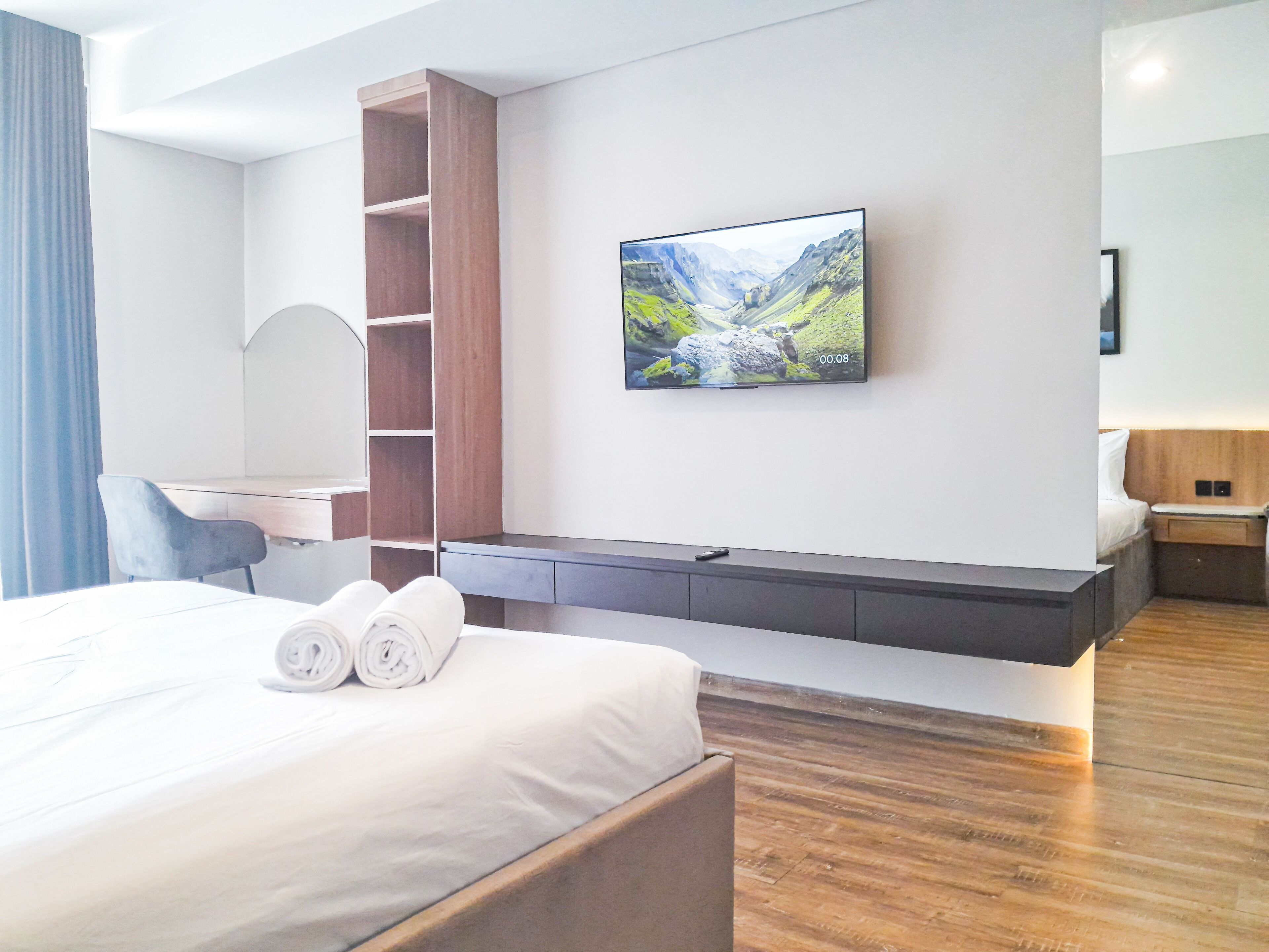 A modern hotel room with a bed, a TV mounted on the wall, a wooden desk, and a built-in shelf.