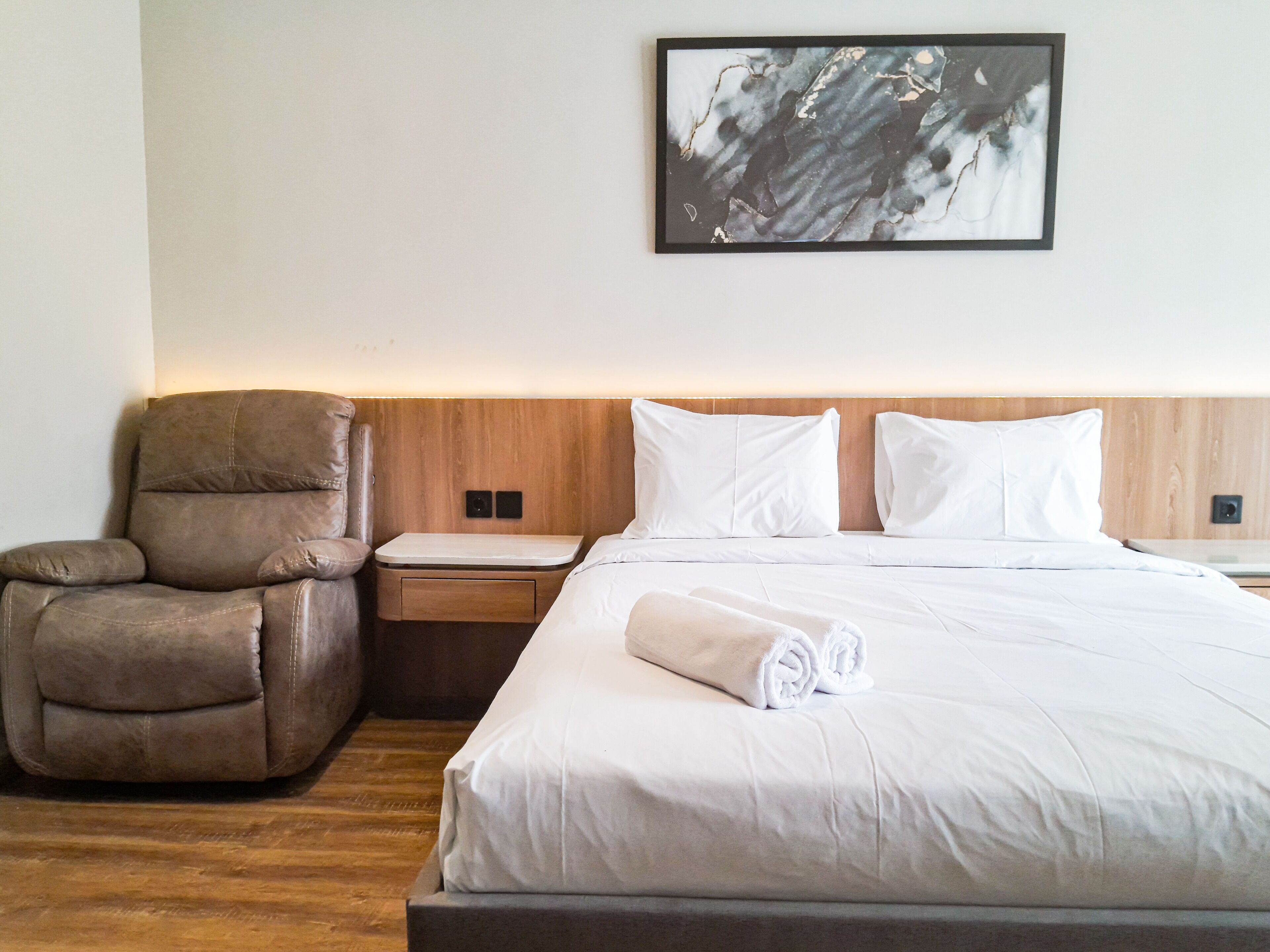 A hotel room with a bed, a chair, a nightstand, and a wall-mounted artwork.
