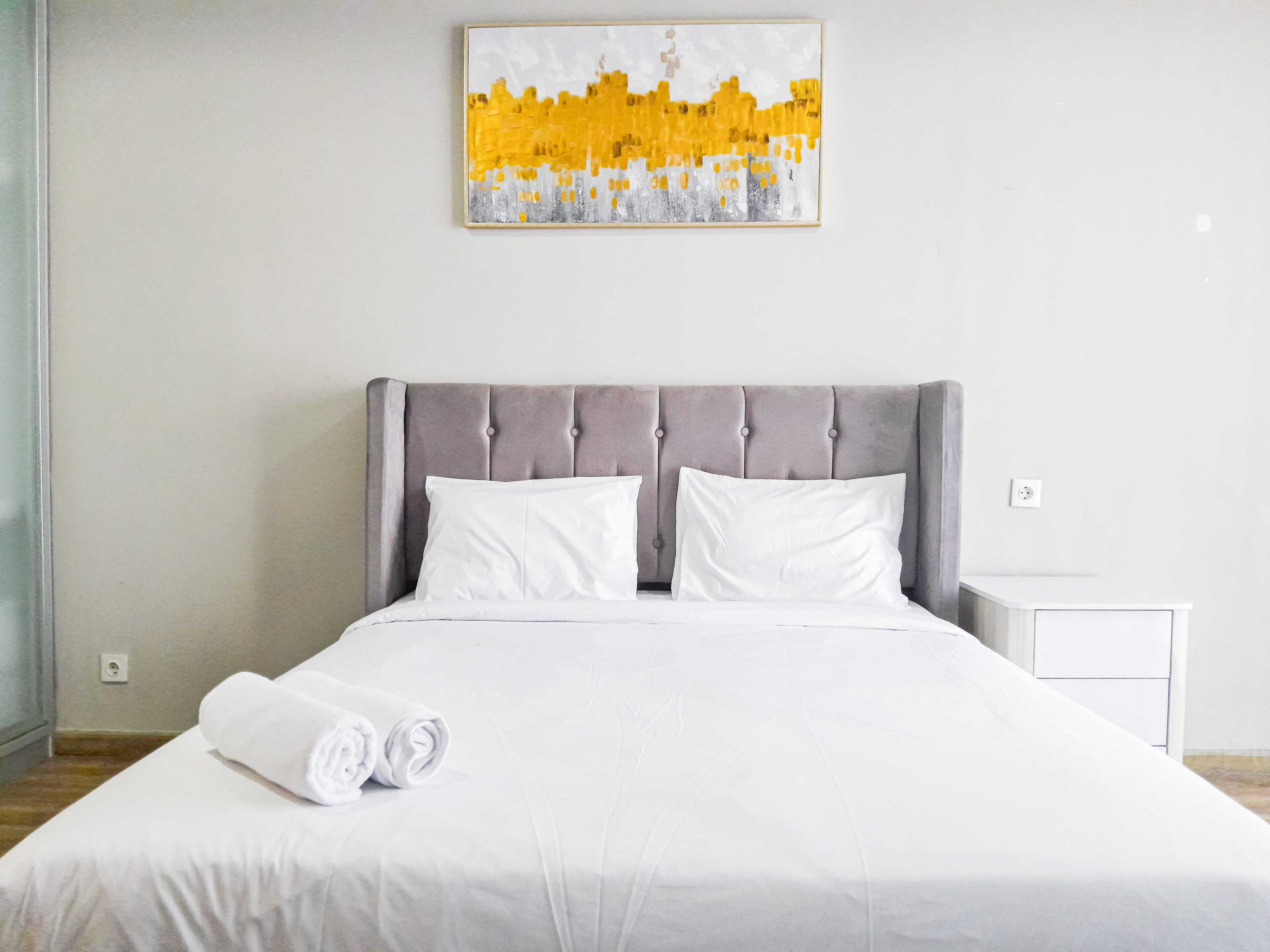 A neatly made bed with a grey upholstered headboard, white pillows, and a rolled white towel on the bed.