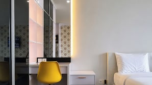 A modern hotel room with a bed, a desk with a chair, a small cabinet, and a large mirror. - Cozy And Chic Studio At Apartment Tamansari Skylounge Balikpapan (Balikpapan)