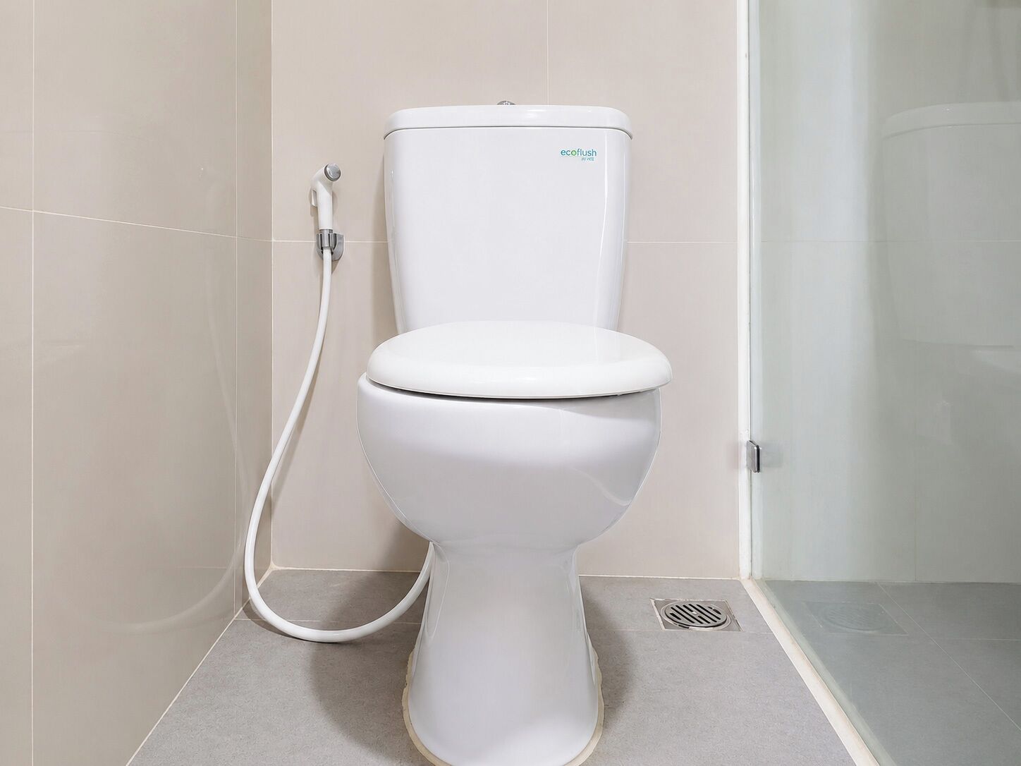 Des toilettes blanches, dotées d’une poignée de chasse d’eau et d’un accessoire de type bidet, dans une salle de bain carrelée de carreaux beiges.