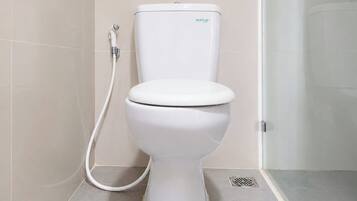 Des toilettes blanches, dotées d’une poignée de chasse d’eau et d’un accessoire de type bidet, dans une salle de bain carrelée de carreaux beiges.