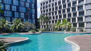 A swimming pool with a wooden deck, surrounded by palm trees and modern buildings. - Functional Nice Studio Room At Apartment Tamansari Skylounge Balikpapan (Balikpapan)