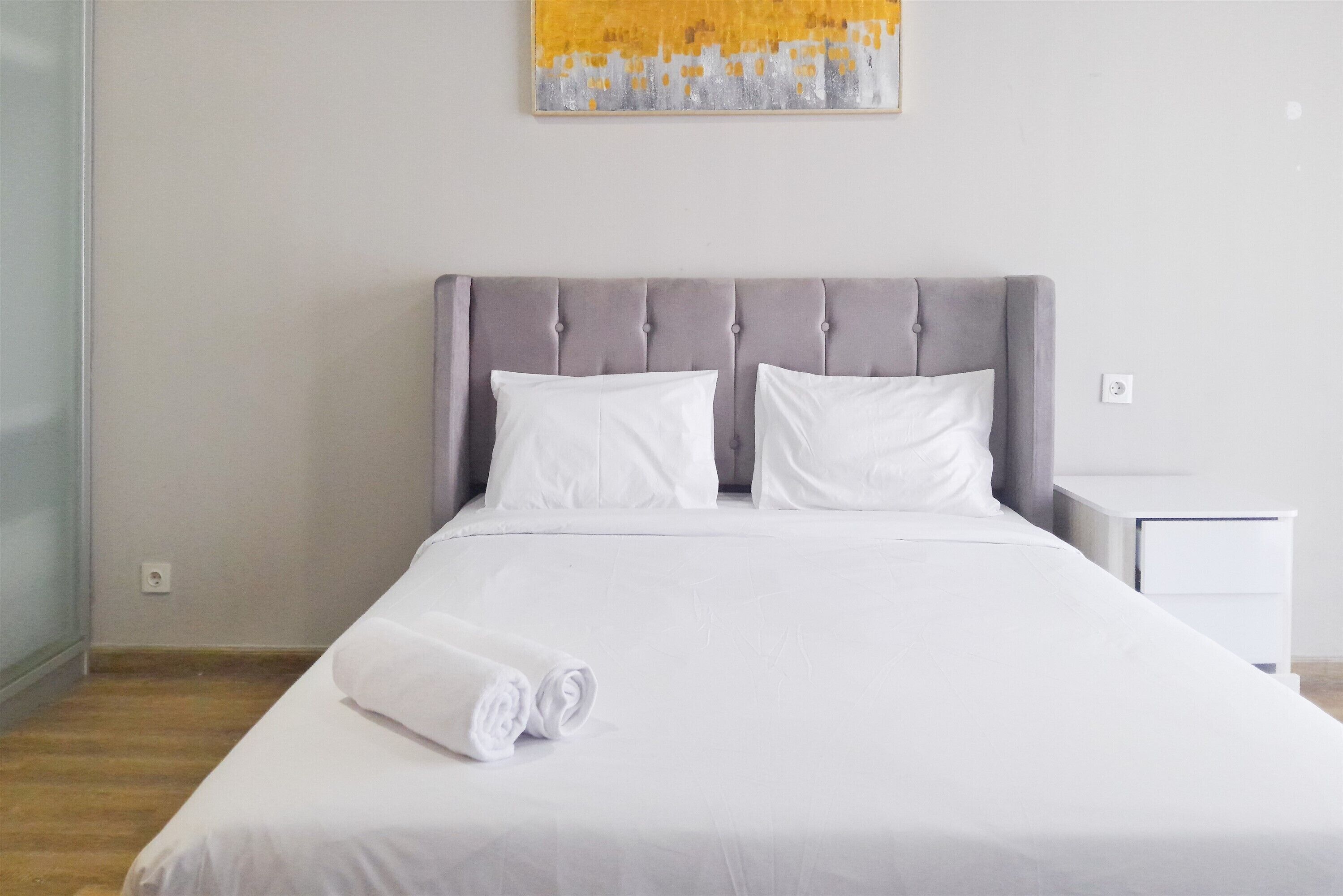 A neatly made bed with a grey upholstered headboard, white bedding, and a rolled white towel on the foot of the bed.