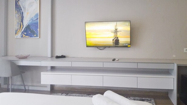 A modern hotel room with a flat-screen TV mounted on the wall, a white console table, a bed with a rolled white towel, and a decorative bowl on the console table. - Functional Nice Studio Room At Apartment Tamansari Skylounge Balikpapan (Balikpapan)