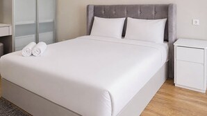 Una cama bien hecha con sábanas blancas y dos toallas enrolladas, un cabecero tapizado en gris y una mesita de noche pequeña de color blanco con un cajón.