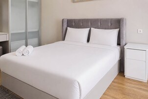 Una cama bien hecha con sábanas blancas y dos toallas enrolladas, un cabecero tapizado en gris y una mesita de noche pequeña de color blanco con un cajón.