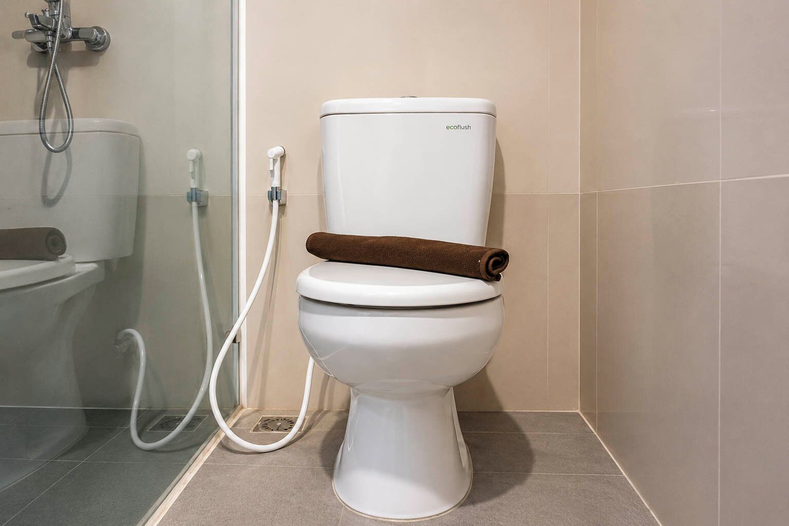 Des toilettes avec un porte-rouleau de papier toilette marron, une cuvette blanche et une pomme de douche visible à l’arrière-plan.