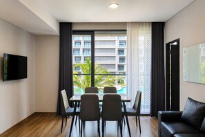 A modern hotel room with a dining table, four chairs, a flat-screen TV, a black leather sofa, and a large window with a view of a building and greenery. - Modern Cozy 1Br Combined Unit At Apartment Tamansari Skylounge Balikpapan (Balikpapan)