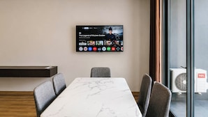 Et moderne spiseområde med et marmorbord, grå polstrede stole, et ophængt TV der viser et program, og udsigt udendørs gennem store vinduer.