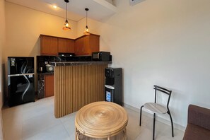 Une cuisine compacte avec des meubles en bois, un four à micro-ondes, une machine à café et un distributeur d’eau. On aperçoit une table ronde en osier et une chaise au premier plan.
