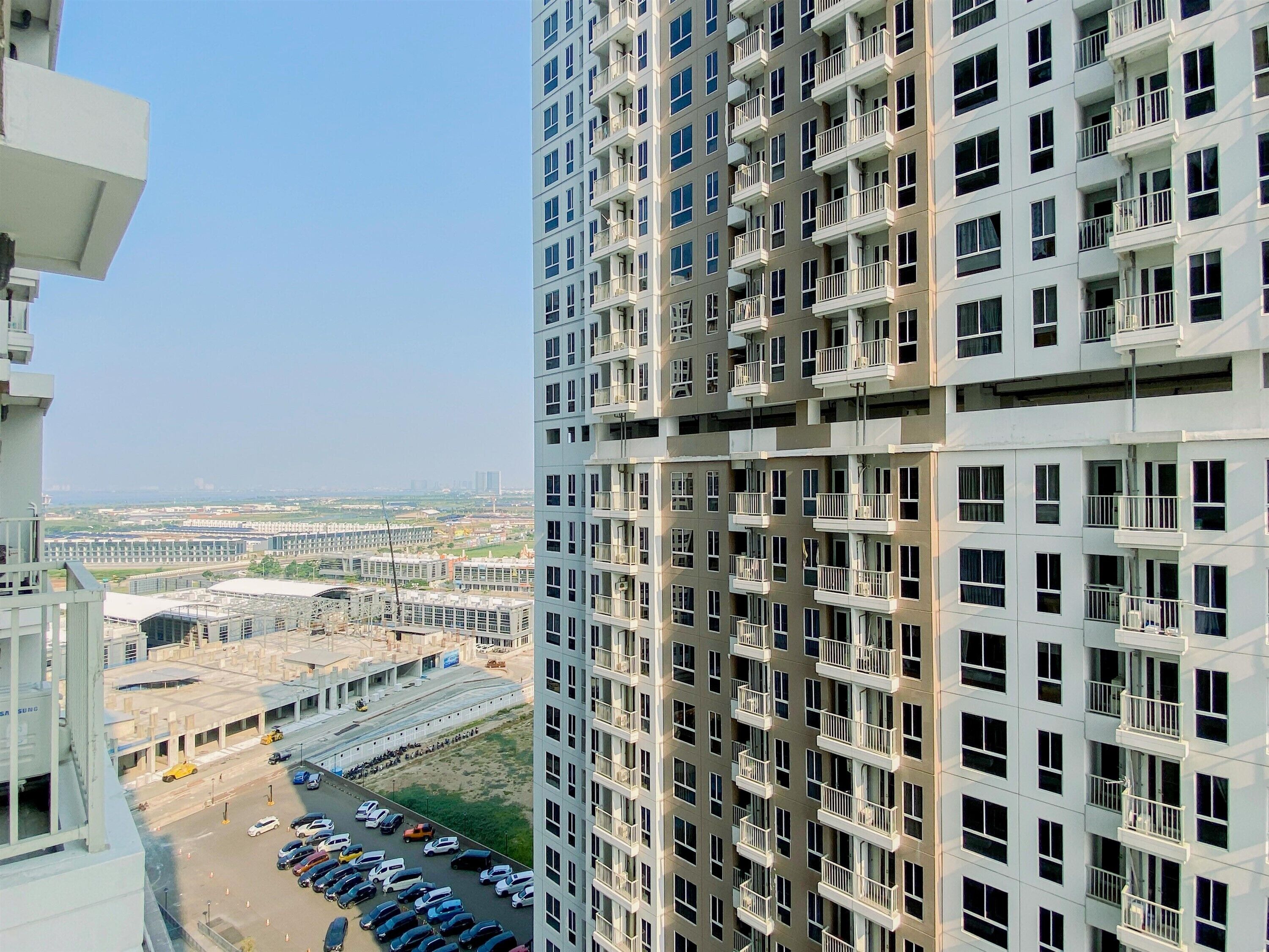 A high-rise building with multiple balconies, a parking lot with cars, and a distant view of a cityscape.