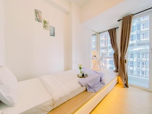 A minimalist bedroom with a single bed, a small table with a vase, and a window with curtains. - Good Place And Homey Studio At Tokyo Riverside Pik 2 Apartment (Jakarta)