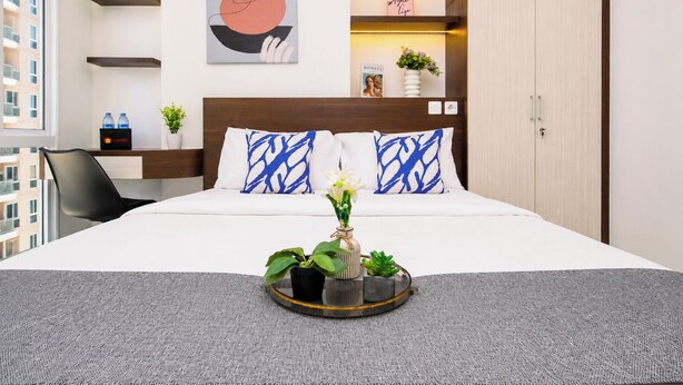 A hotel room with a bed, a bedside table, a chair, a window, and a wall with a painting and a shelf with decorative items. - Studio Polished Areum Residence At Tokyo Riverside (Jakarta)
