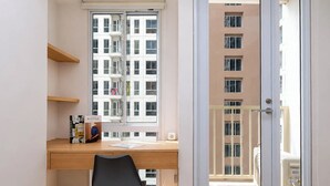 Un appartement moderne doté d’un bureau en bois, d’une chaise noire, d’une fenêtre donnant sur un immeuble et d’un balcon avec un ventilateur.