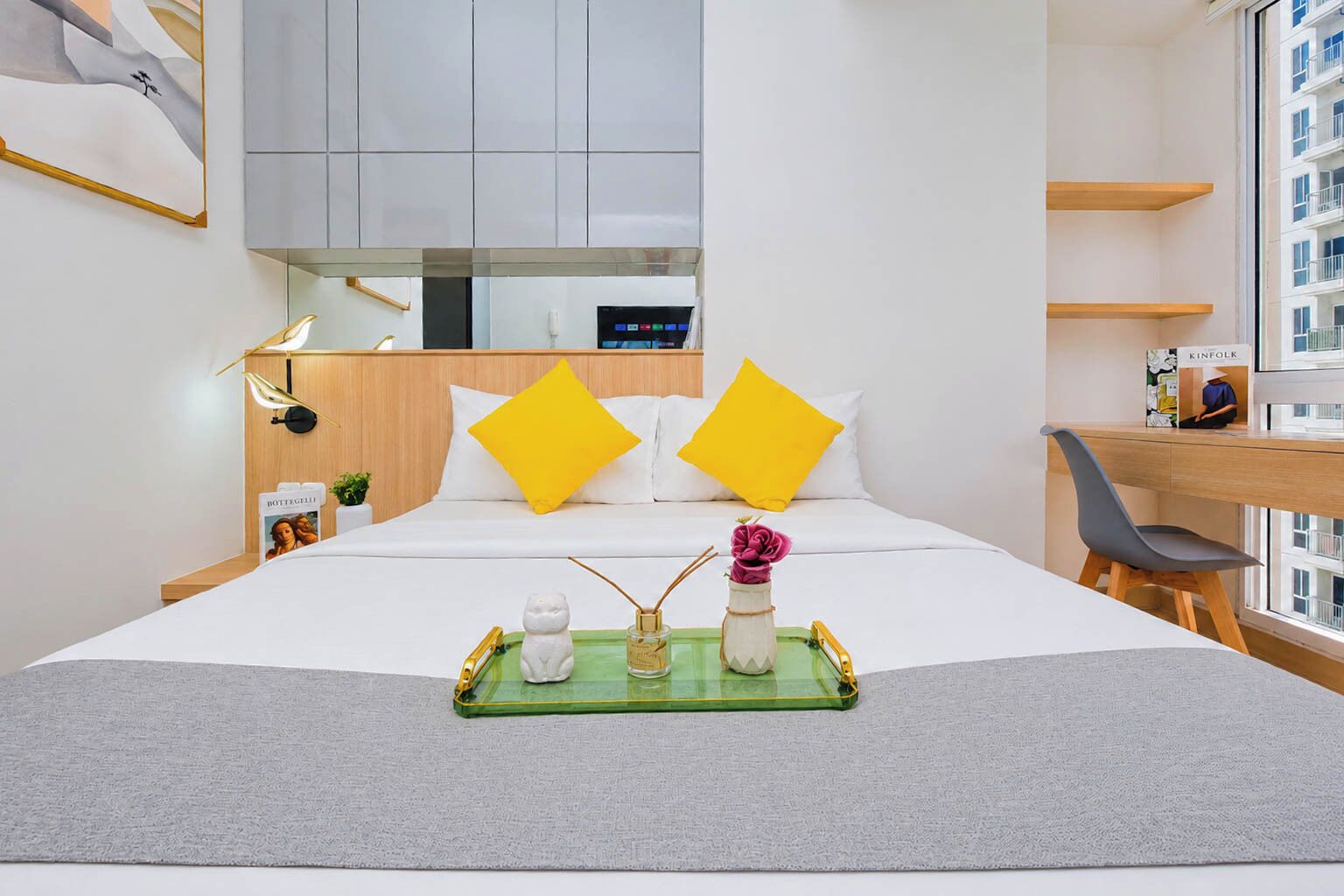 A hotel room with a bed, a desk, a chair, a window with a view of buildings, and a tray with items on the bed.