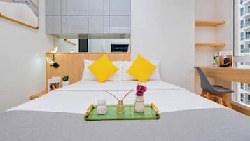 A hotel room with a bed, a desk, a chair, a window with a view of buildings, and a tray with items on the bed.