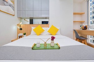 Habitación de hotel con cama, escritorio, silla, ventana con vista a edificios y una bandeja con objetos sobre la cama.