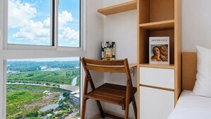 A room with a view of a river and greenery, a wooden chair, a bookshelf with a book, and a bed with white bedding. - Clean And Warm Studio Room At Apartment Tokyo Riverside Pik 2 (Teluknaga)