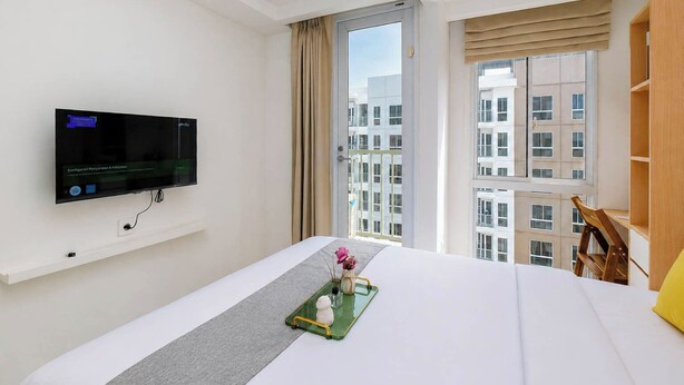 A hotel room with a bed, a TV mounted on the wall, a window with a view of buildings, and a wooden shelf. - Clean And Warm Studio Room At Apartment Tokyo Riverside Pik 2 (Teluknaga)