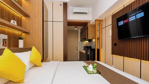 Quarto de hotel moderno com cama, televisão exibindo serviços de streaming, cozinha compacta e banheiro.