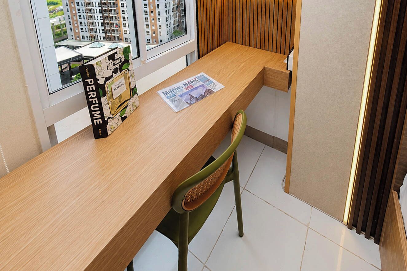A wooden desk with a magazine, a perfume box, and a chair in a modern room with a window and a view of buildings.