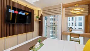 Quarto de hotel com televisão exibindo serviços de streaming, varanda com vista para edifícios, cama com um apoio de mesa verde e escrivaninha com cadeira.