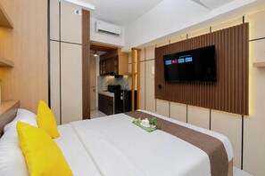 A hotel room with a bed, a TV mounted on the wall, a kitchenette, and an air conditioning unit. - Warm And Homey Studio At Apartment Tokyo Riverside Pik 2 (Jakarta)