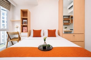 Ein modernes Hotelzimmer mit Bett, orangefarbener Bettwäsche, einem kleinen Tisch mit Pflanze, einer Bücherwand und einem Fenster mit Blick auf Gebäude.