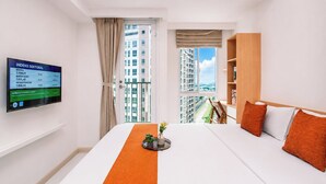 A hotel room with a bed, a television displaying an index, a window with a view of buildings, and a wooden shelf. - Good Deal And Minimalist Studio Apartment At Tokyo Riverside Pik 2 (Jakarta)