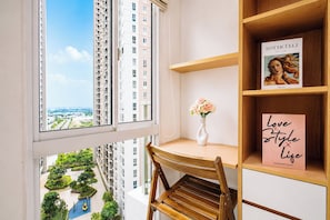 A corner room with a view of high-rise buildings, a wooden chair, a white vase with flowers, and a shelf with a framed picture and a sign. - Good Deal And Minimalist Studio Apartment At Tokyo Riverside Pik 2 (Jakarta)