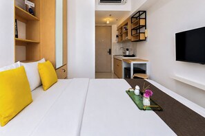 A hotel room with a bed, a small kitchenette, a TV mounted on the wall, and a shelf with books. - Good Deal And Homey Studio Apartment Tokyo Riverside Pik 2 (Jakarta)