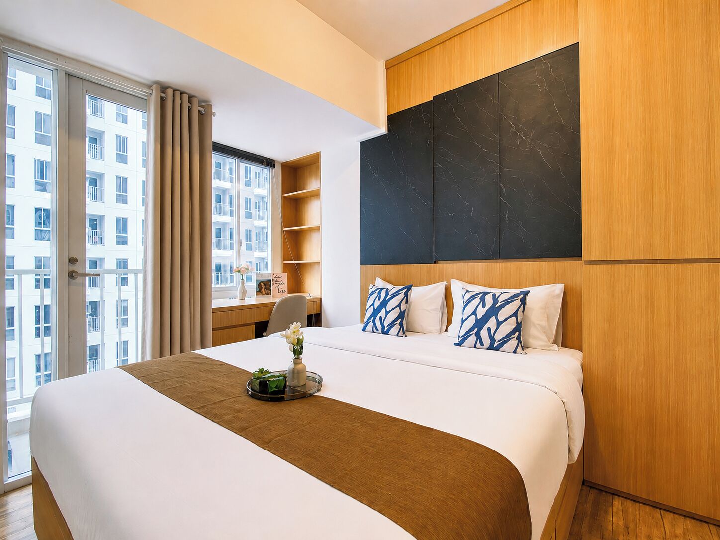 A hotel room with a large bed, a wooden headboard, a view of the city through the window, and a desk with a chair.