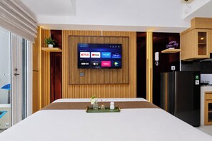 Une chambre d’hôtel moderne dotée d’un grand téléviseur à écran plat fixé sur un mur en panneaux de bois, d’un lit avec une tête de lit blanche, d’une petite table avec un vase et d’un réfrigérateur.