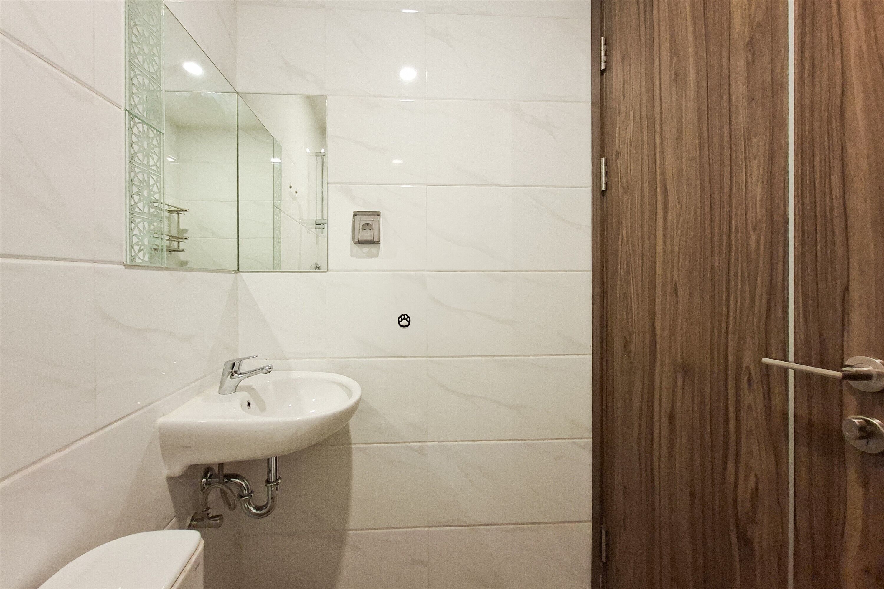 Baño con lavamanos blanco, espejo, inodoro y puerta de madera, además de paredes con azulejos.
